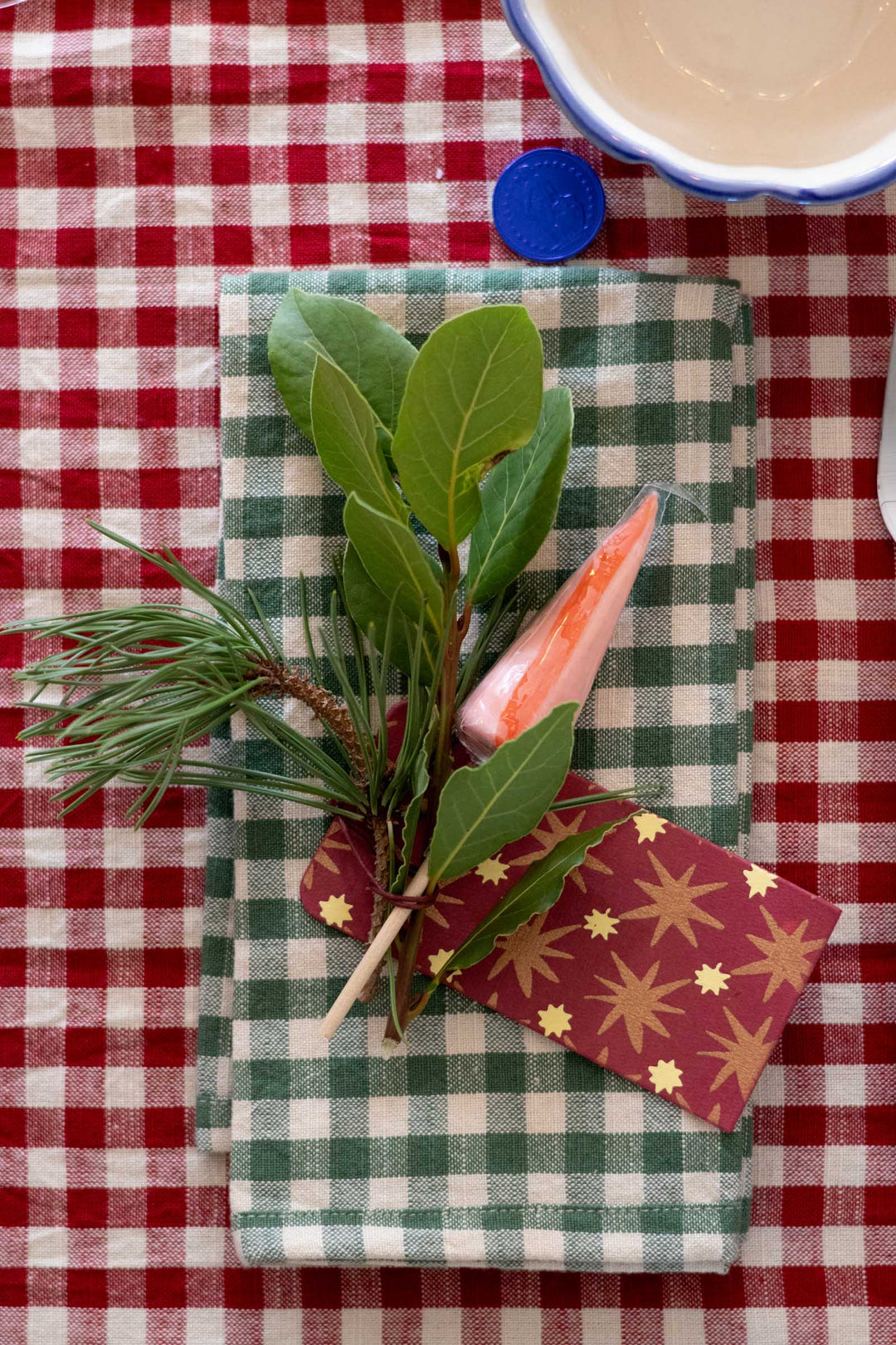 A green and white checkered cloth napkin wrapped with a pine sprig, bay leaf, and a pink decorative paper accent, laid on a red and white checkered tablecloth.