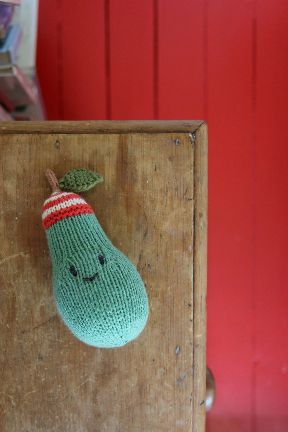 Knitted green eggplant with a red and white striped hat on a wooden surface with a red background