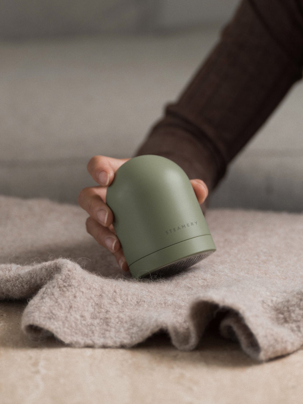 Person holding a green fabric shaver with 'steamery' branding on a textured surface.