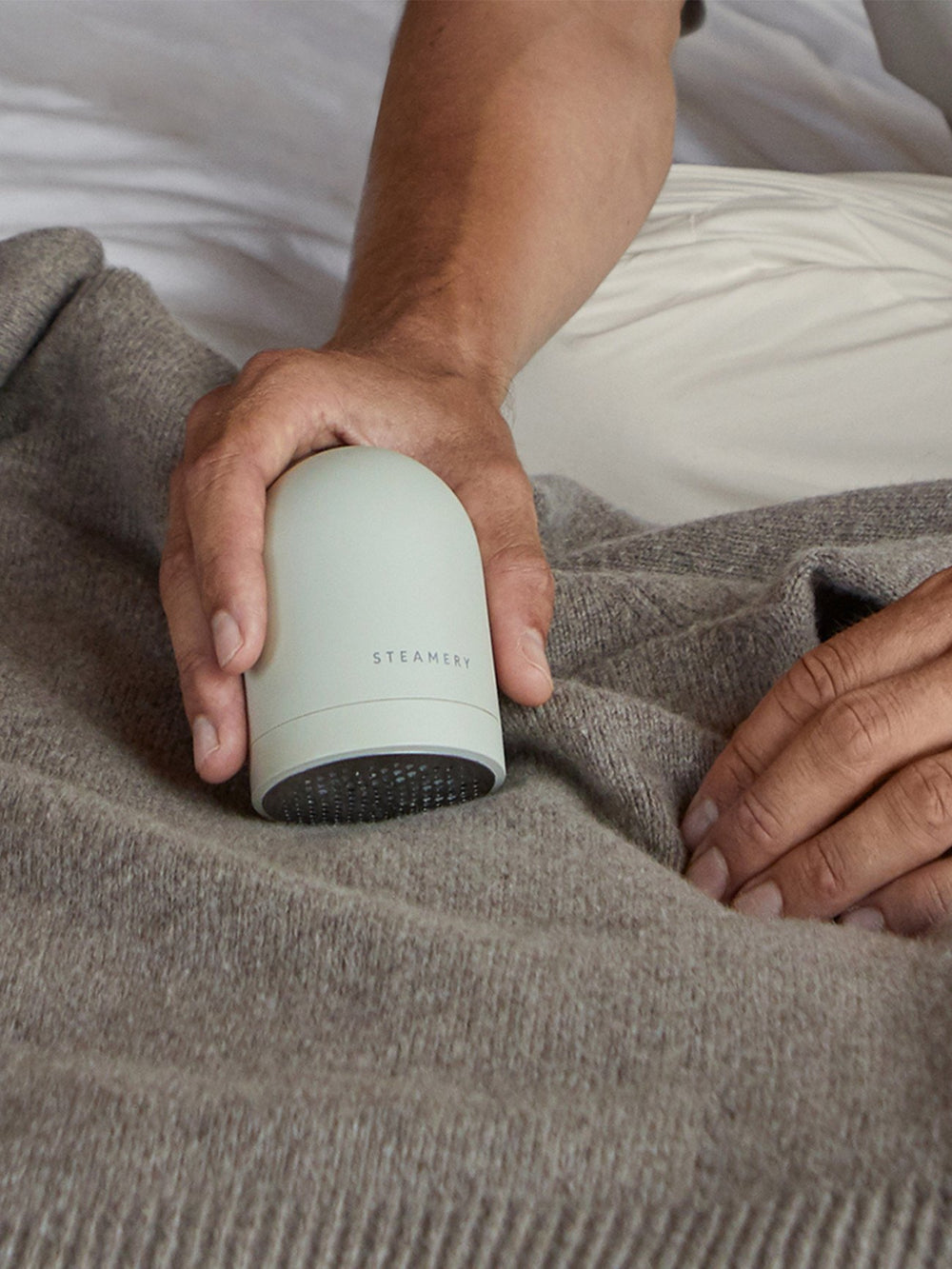 Person holding a small cylindrical fabric shaver labeled 'Steamery' under a blanket