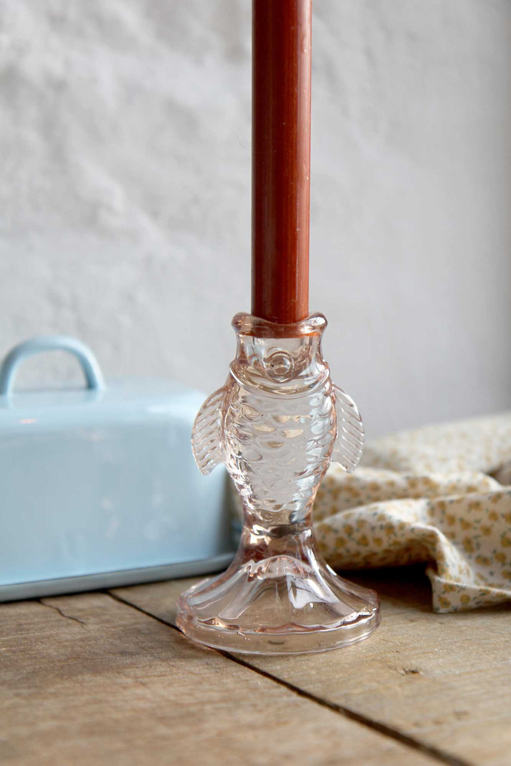 Clear glass candle holder with a red candle on a wooden surface with a light blue container and patterned fabric in the background.