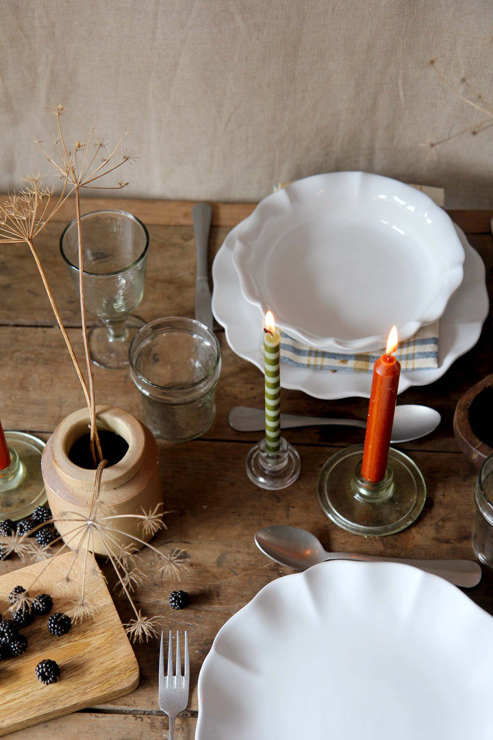 Dining table setting with white plates, glasses, candles, and cutlery on a wooden surface.