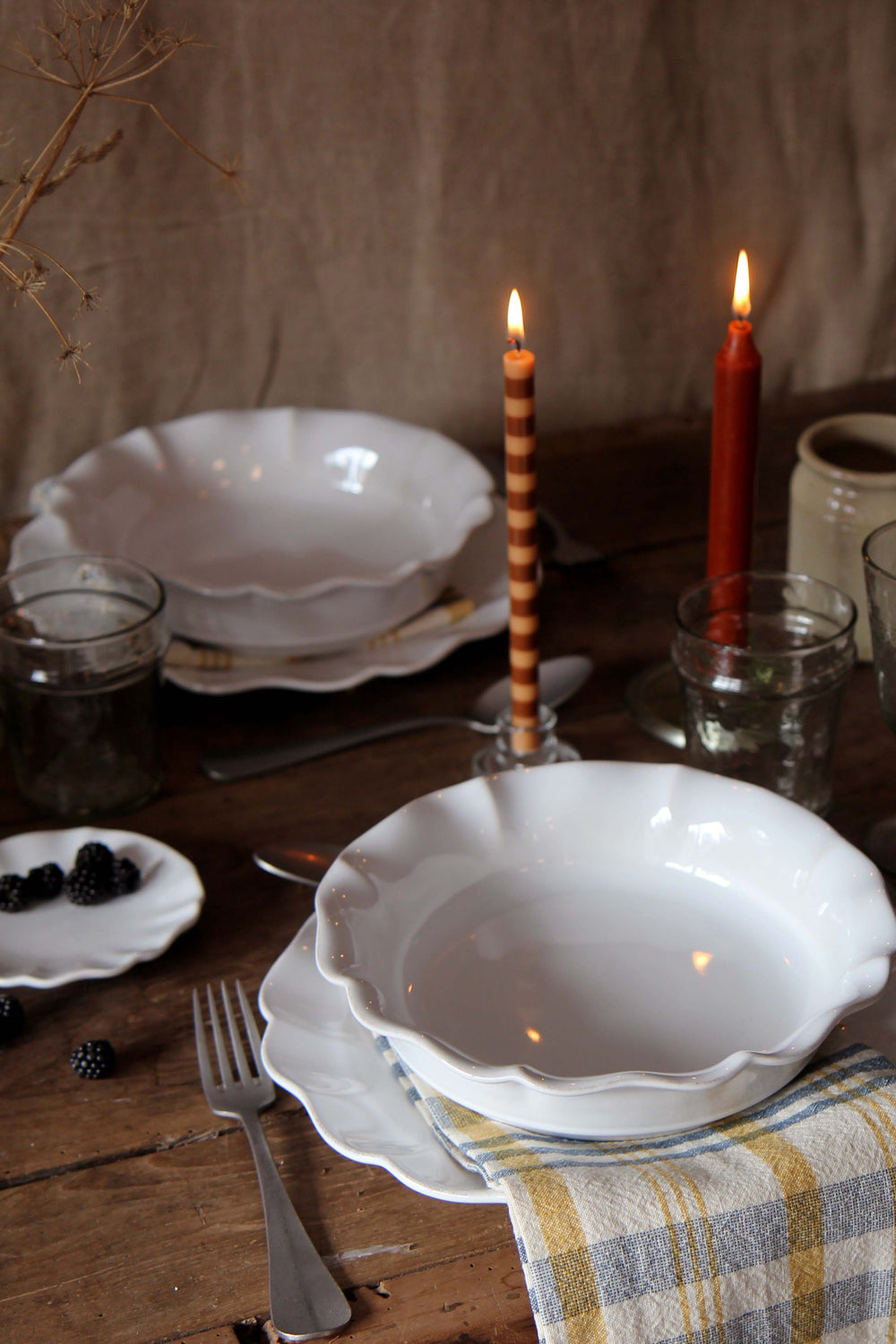 Set dining table with white pasta bowls, candles, and cutlery on a wooden surface.