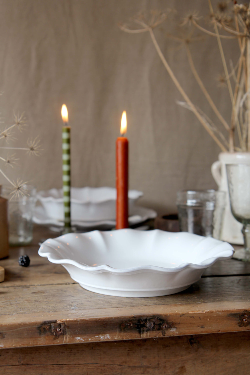 White scalloped-edge soup plate on a wooden surface with lit candles and decorative elements.