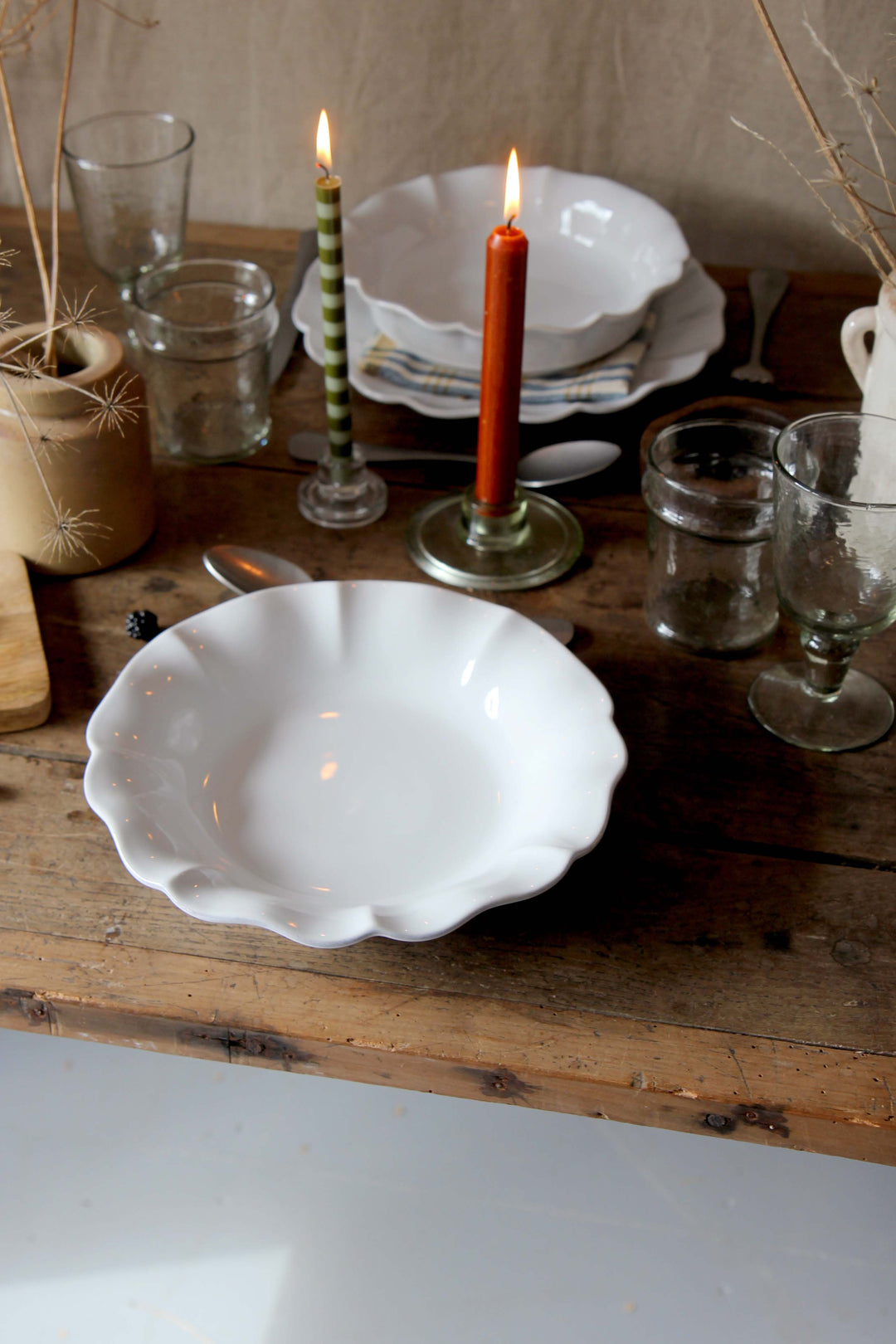 Dining table setup with white soup plate, glasses, and lit candles on a wooden surface.