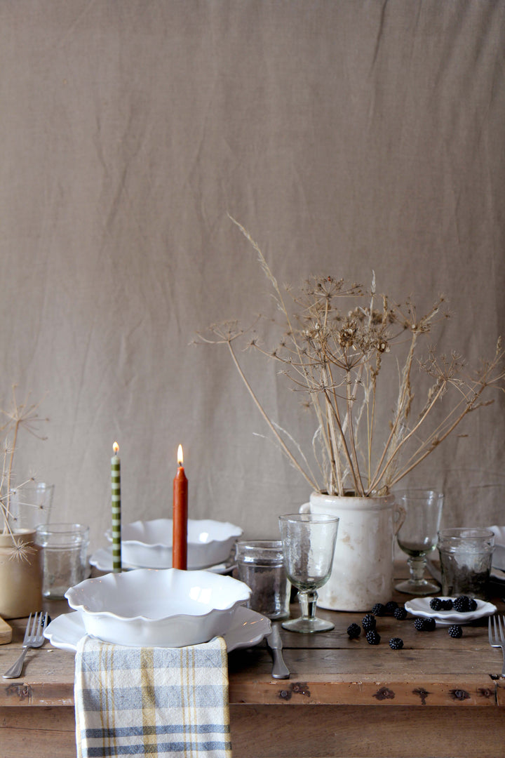 Dining table setting with rosa white scalloped tableware, glasses, and a vase on a wooden surface.