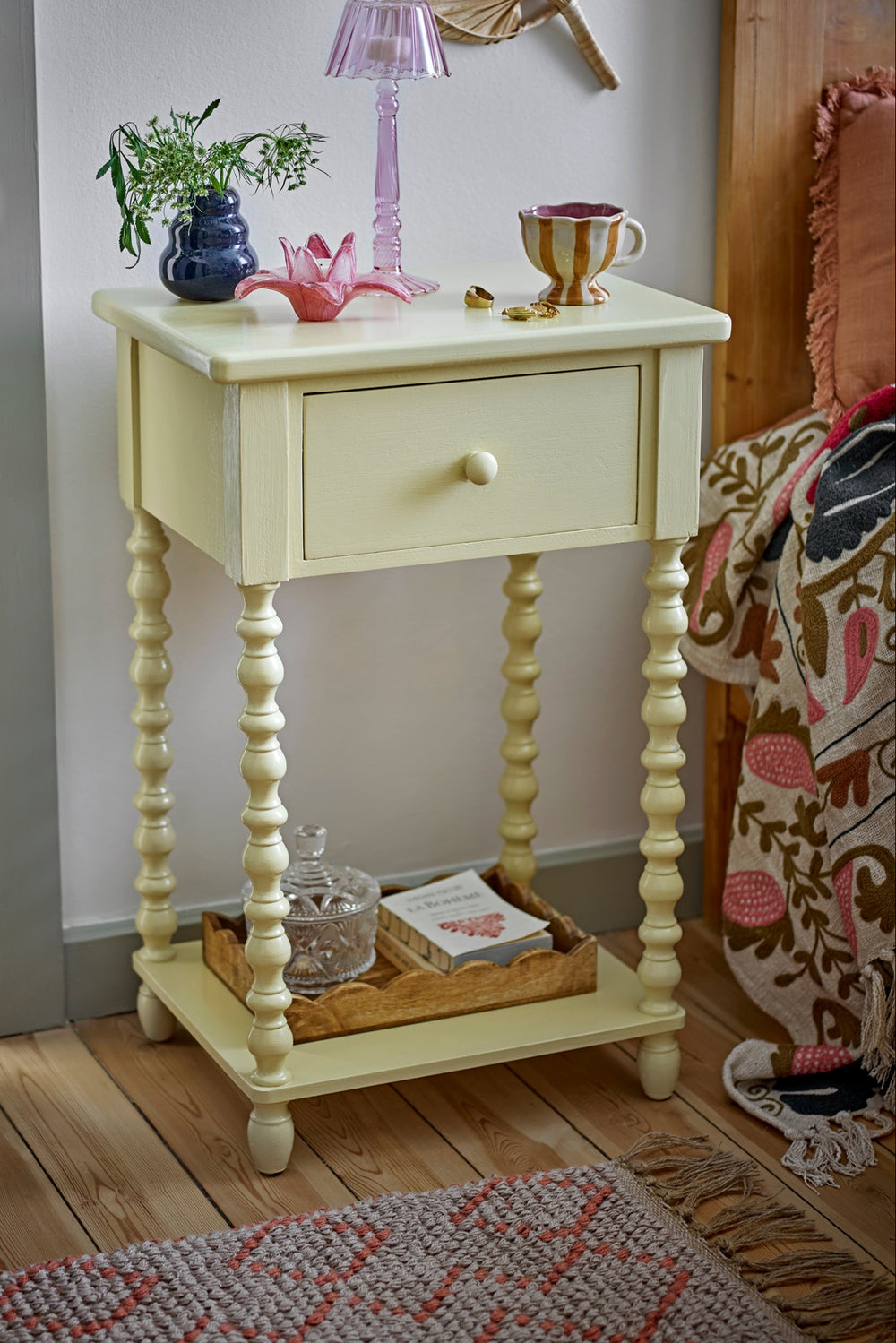 Small cream-colored side table with a lamp, books, and decorative items in a room.