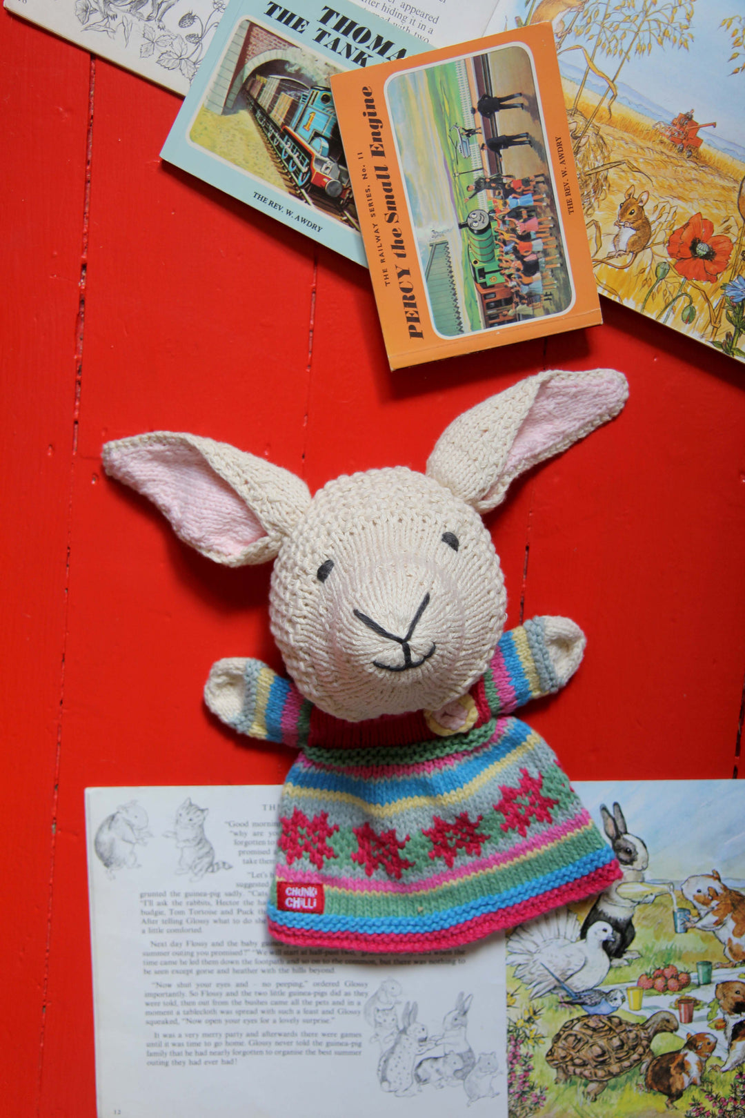 Knitted sheep toy with a colourful sweater on a red surface with vintage books and illustrations.