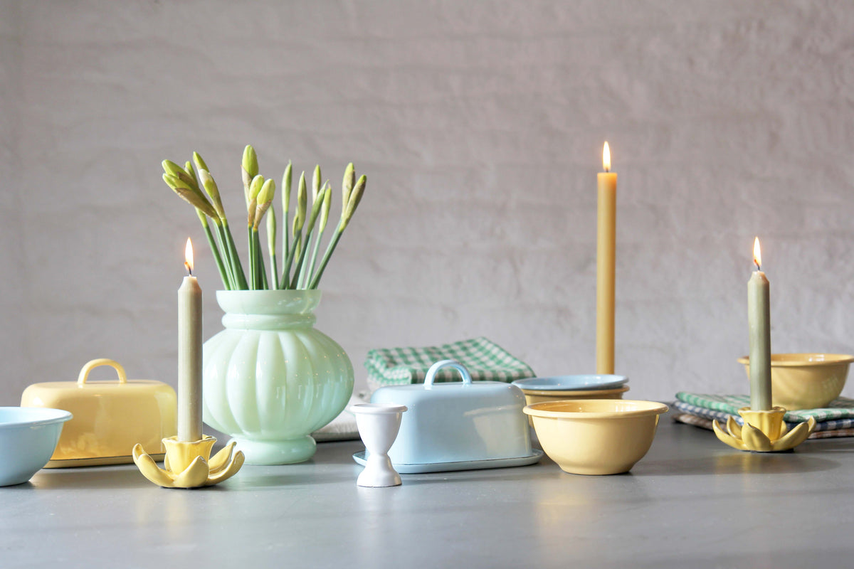 table with spring coloured tableware and enamel in blues and yellows. Green vase with daffodils and lit dinner candle.