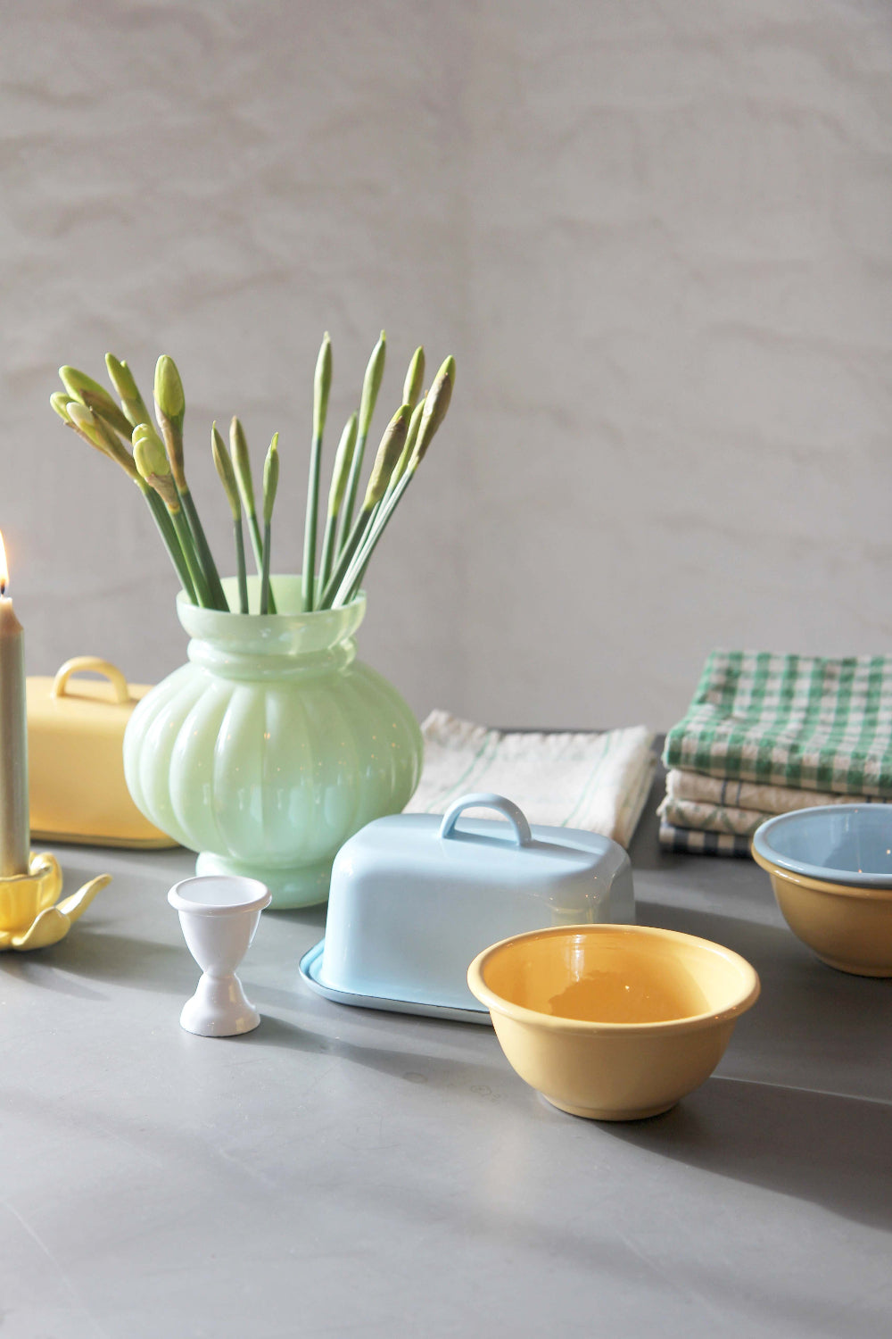 table with spring coloured tableware and enamel in blues and yellows. Green vase with daffodils and lit dinner candle.