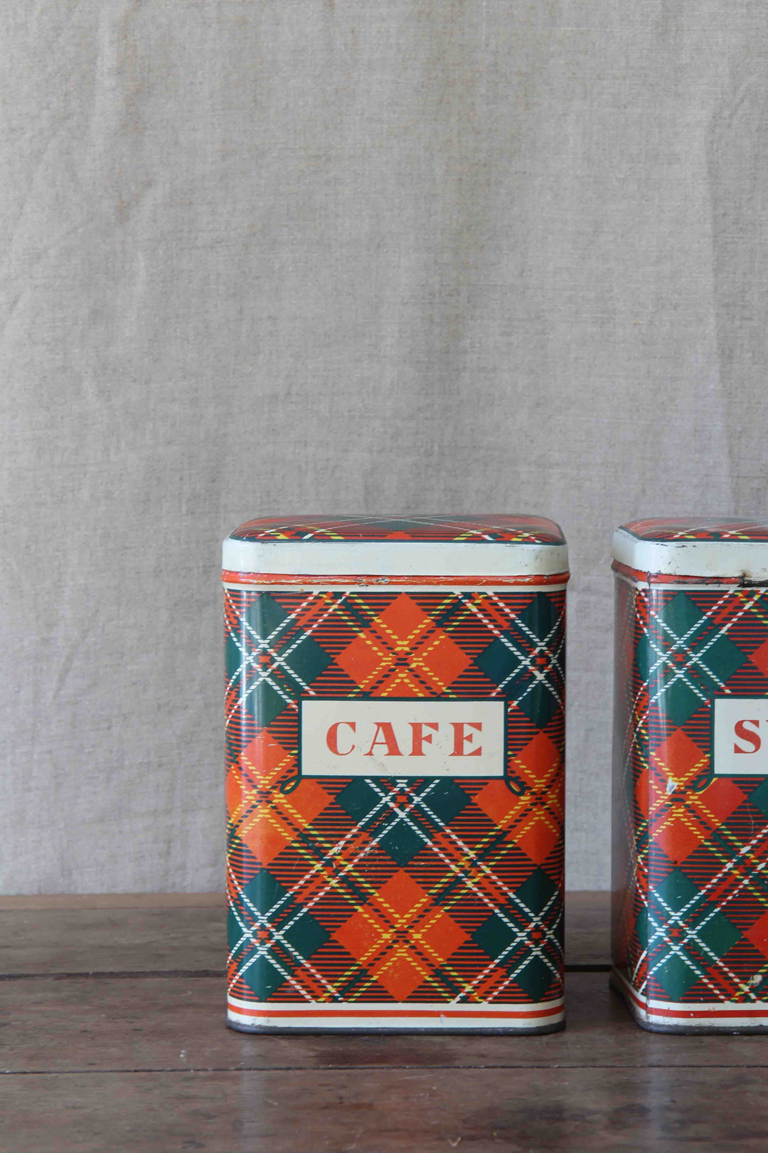 Two vintage-style metal canisters with plaid patterns and 'CAFE' labels on a wooden surface.