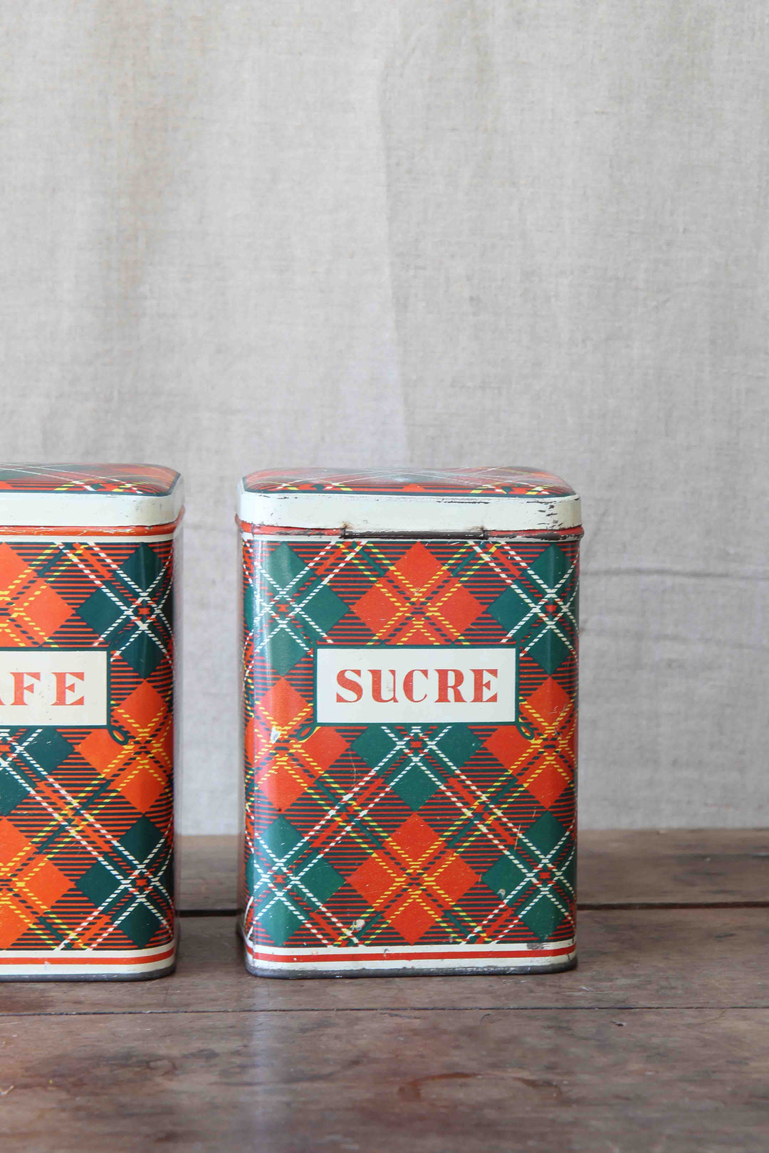 Two plaid-patterned tins labeled 'Sucre' on a wooden surface with a textured gray background.