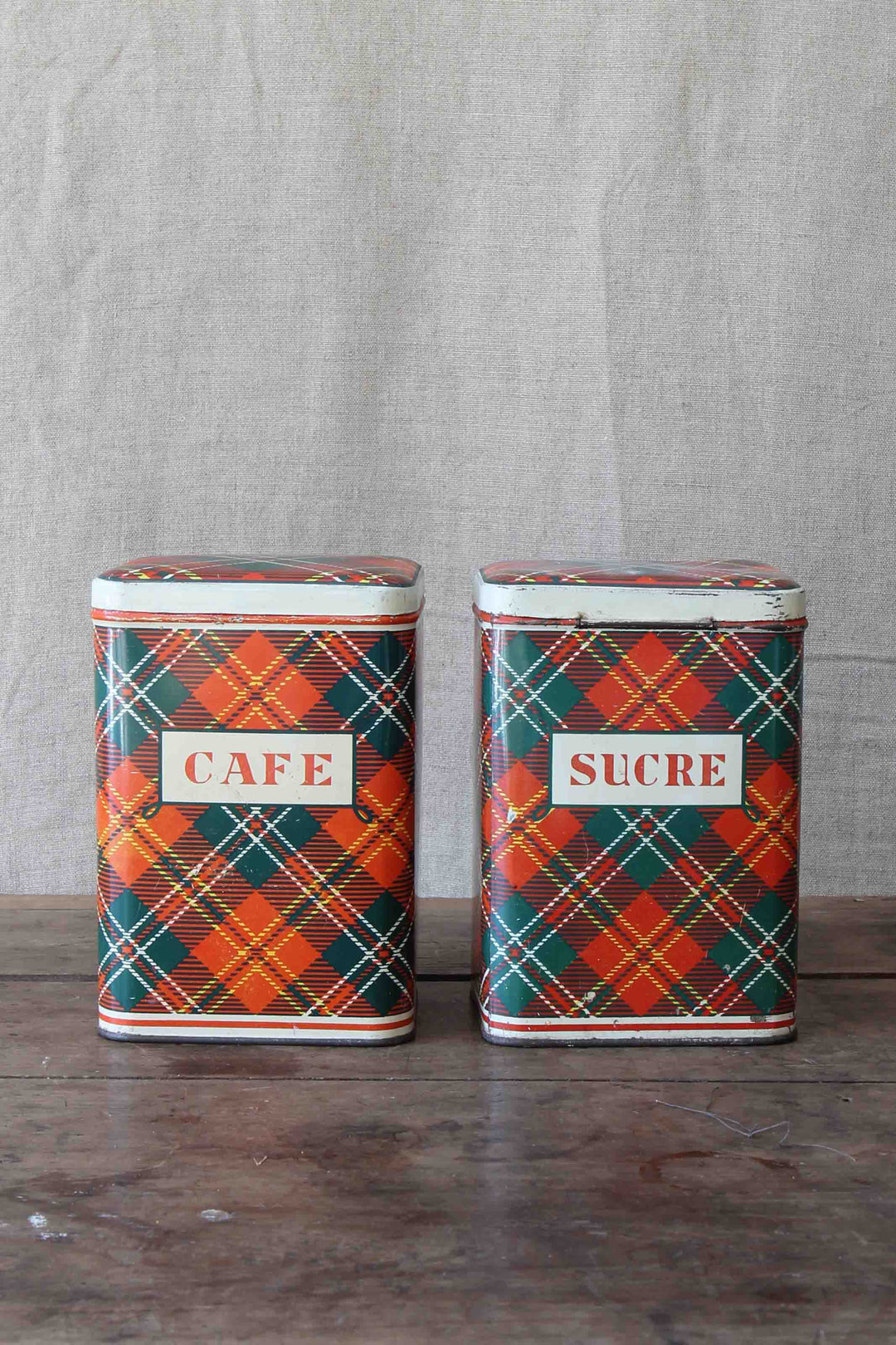 Two plaid-patterned tins labeled 'Cafe' and 'Sucre' on a wooden surface with a gray fabric background.