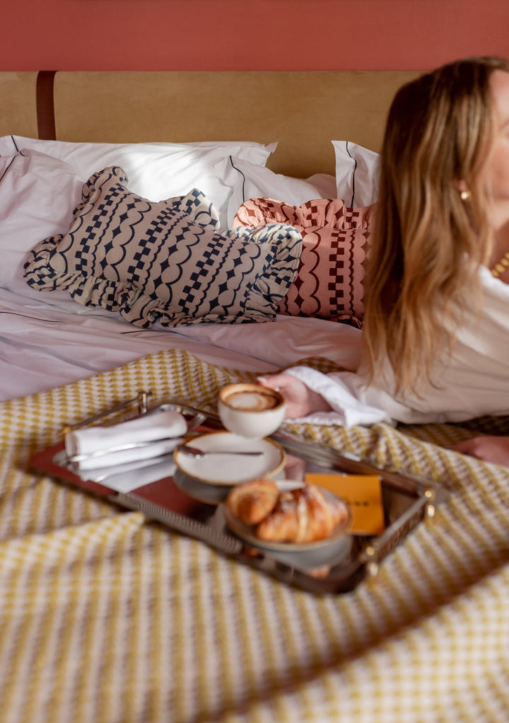 navy geometric patterned oblong cushion with ruffle trim on bed with woman having breakfast