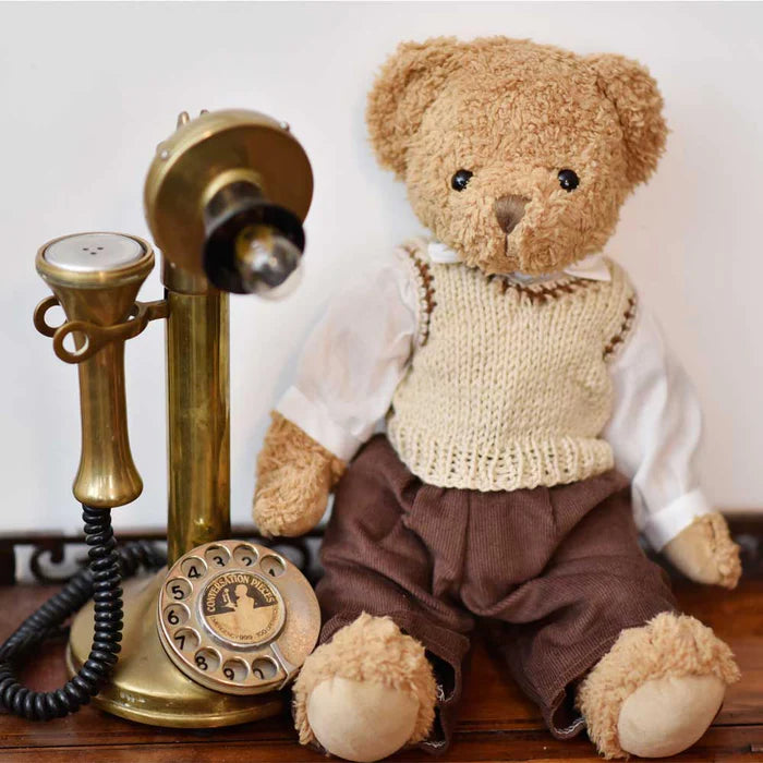Teddy bear in a sweater and pants next to an old-fashioned brass telephone on a wooden surface.