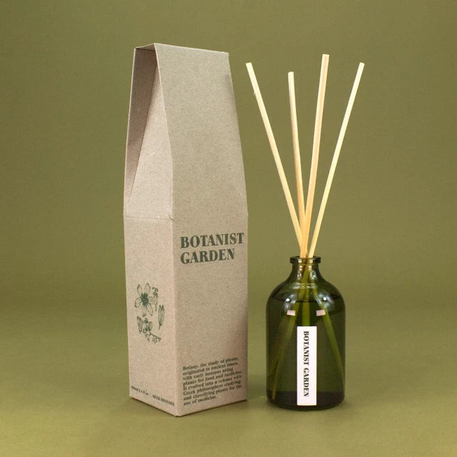 A reed diffuser set with a dark green glass bottle, natural wood reeds, and a tall kraft paper box, all branded Botanist Garden, set against a solid olive green background.