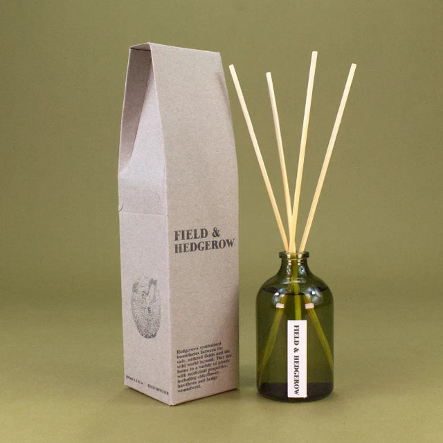 Field & Hedgerow reed diffuser set featuring a dark green glass bottle with natural wooden reeds and its textured kraft paper packaging box, set against a solid olive green background.