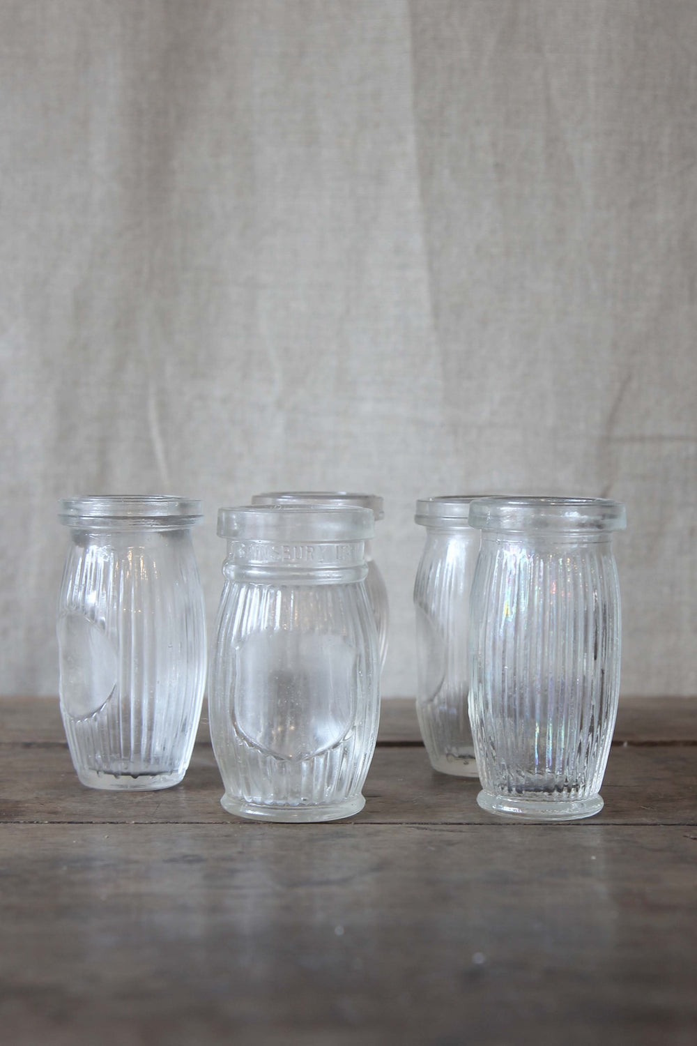 Four clear glass vintage paste jars with ribbed design on a wooden surface and fabric background