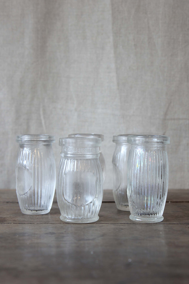 Four clear glass vintage paste jars with ribbed design on a wooden surface and fabric background