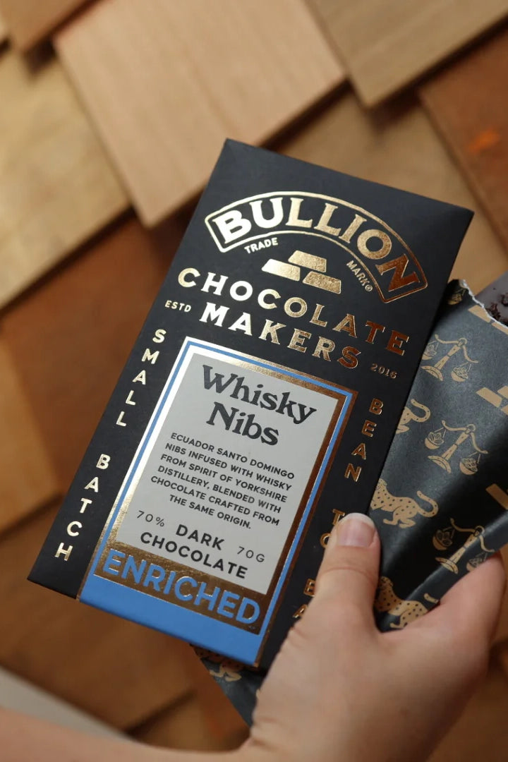 Bullion Chocolate Makers Whisky Nibs chocolate bar packaging held by a hand on a wooden surface.