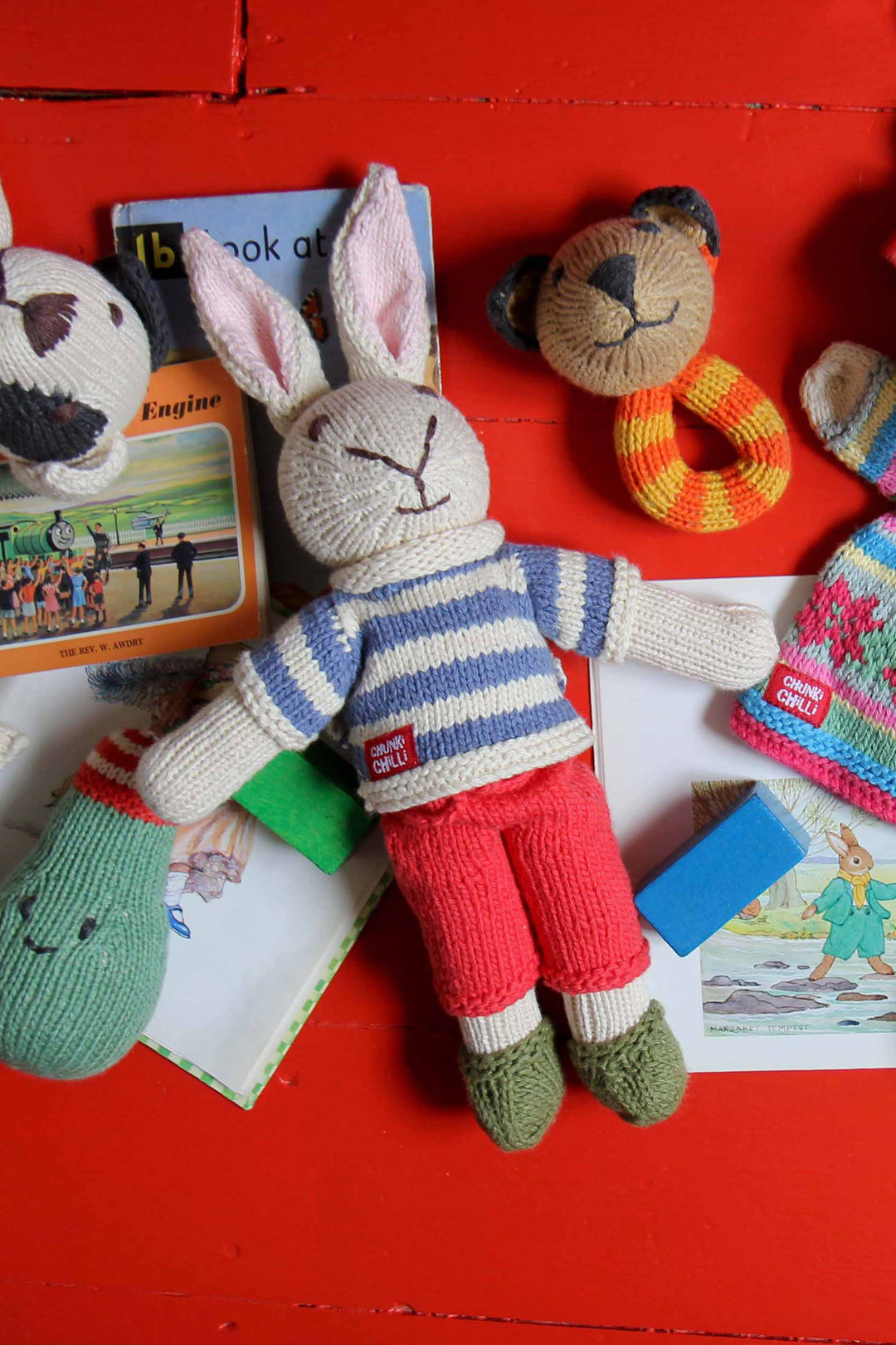 Knitted rabbit toy with a striped sweater on a red surface with books and other toys.