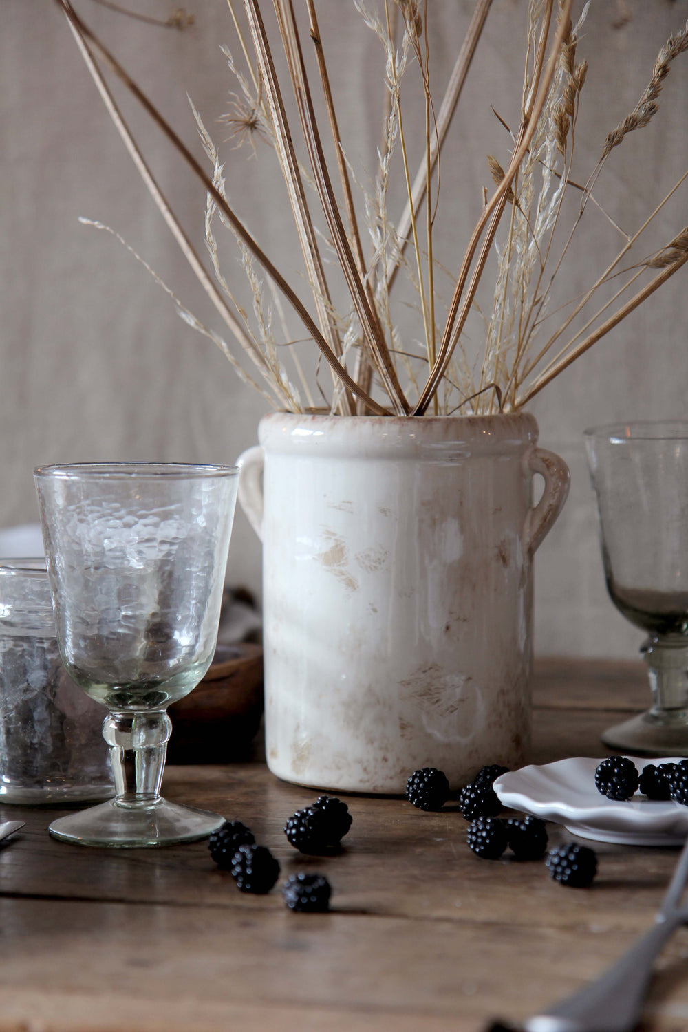 Decorative setting with a ceramic glazed vase, wine glasses, and blackberries on a wooden surface.