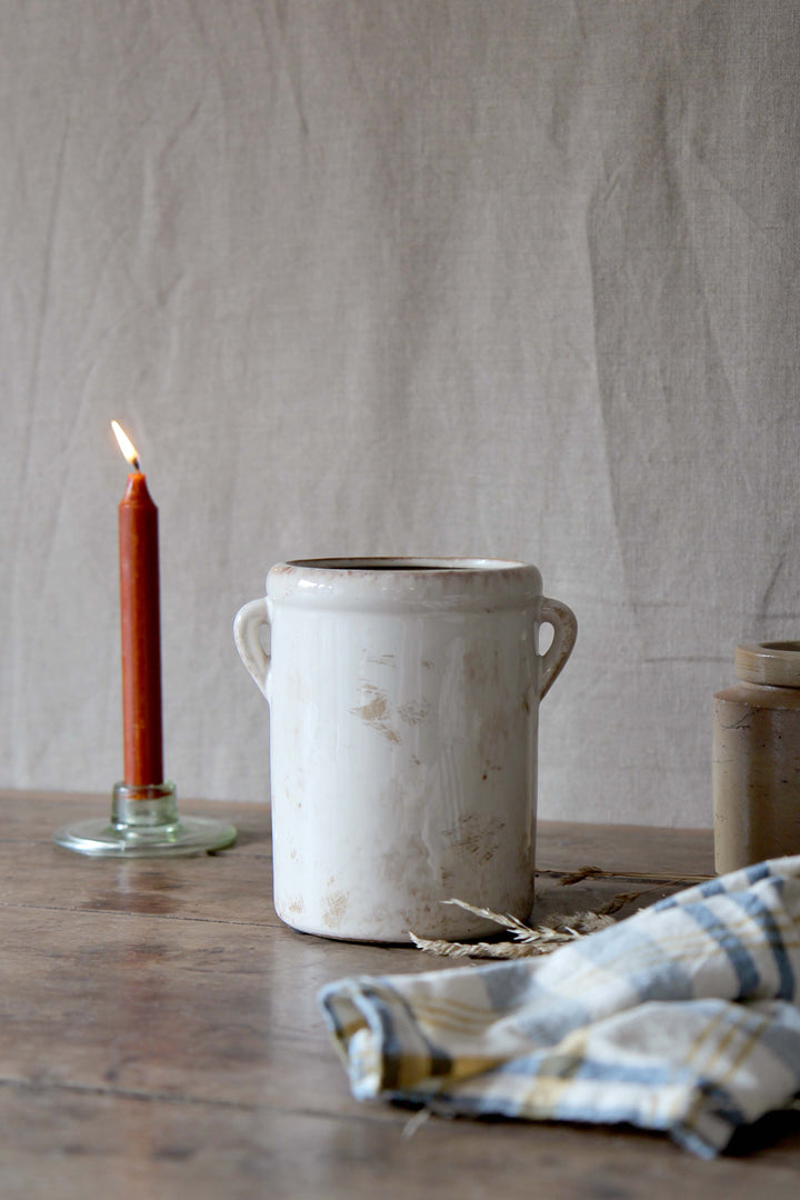 White ceramic jar on a wooden surface with a lit candle and plaid cloth in the background.